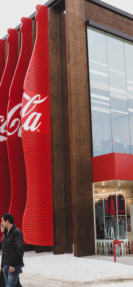 Außenansicht eines rechteckigen Holzpavillons. An der Vorderseite eine große Glasfassade mit der Aufschrift "Coca-Cola", seitlich gehen rote Lamellen über die gesamte Gebäudehöhe. | © LignoAlp