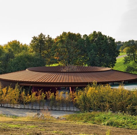Ein ringförmiges Gebäude mit einem Dach aus rostigem Stahl inmitten grüner Landschaft | © Matteo Piazza