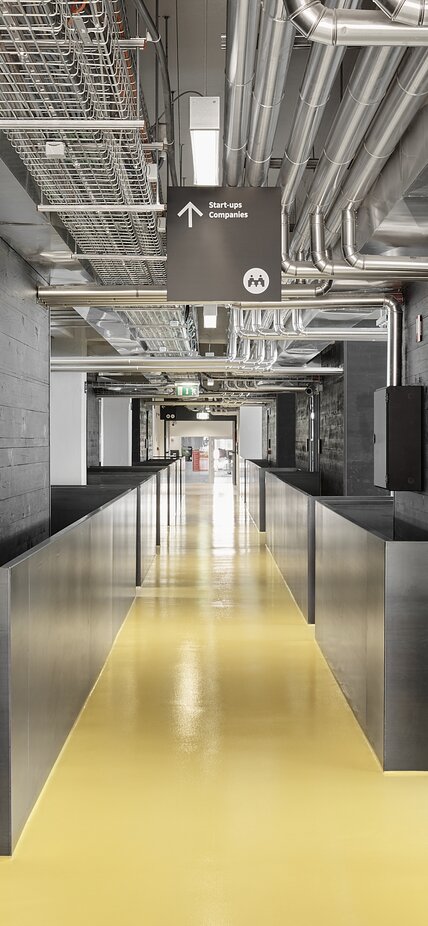 A corridor with a yellow floor leads into a modern-looking building, to the left and right of which are walls made of black impregnated wood | © Davide Perbellini