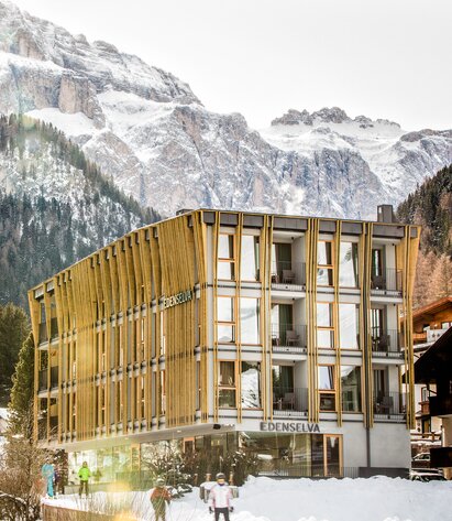 Ein vierstöckiges Hotelgebäude im Winter, die drei Obergeschosse sind mit vertikalen Lamellen verkleidet, im Hintergrund ein Wald und Berge | © Edenselva - Mattia Gasparotto