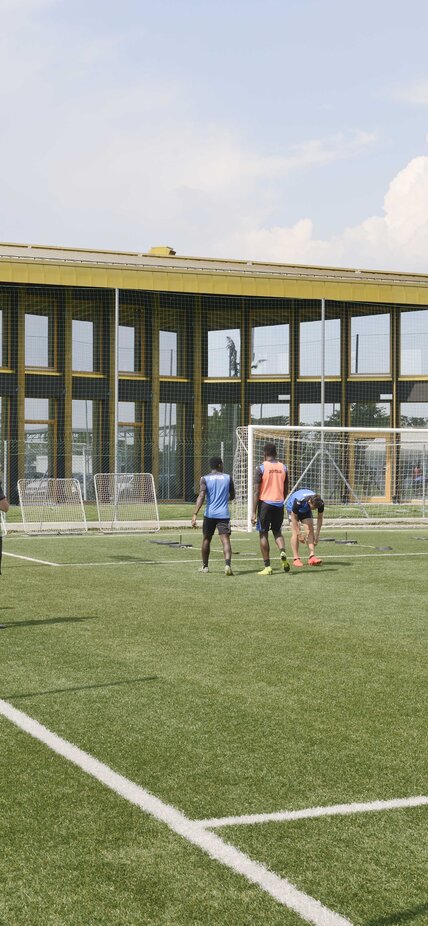 Un campo da calcio con giocatori, sullo sfondo un grande edificio con molte finestre e un tetto color oro lucido | © Michele Nastasi