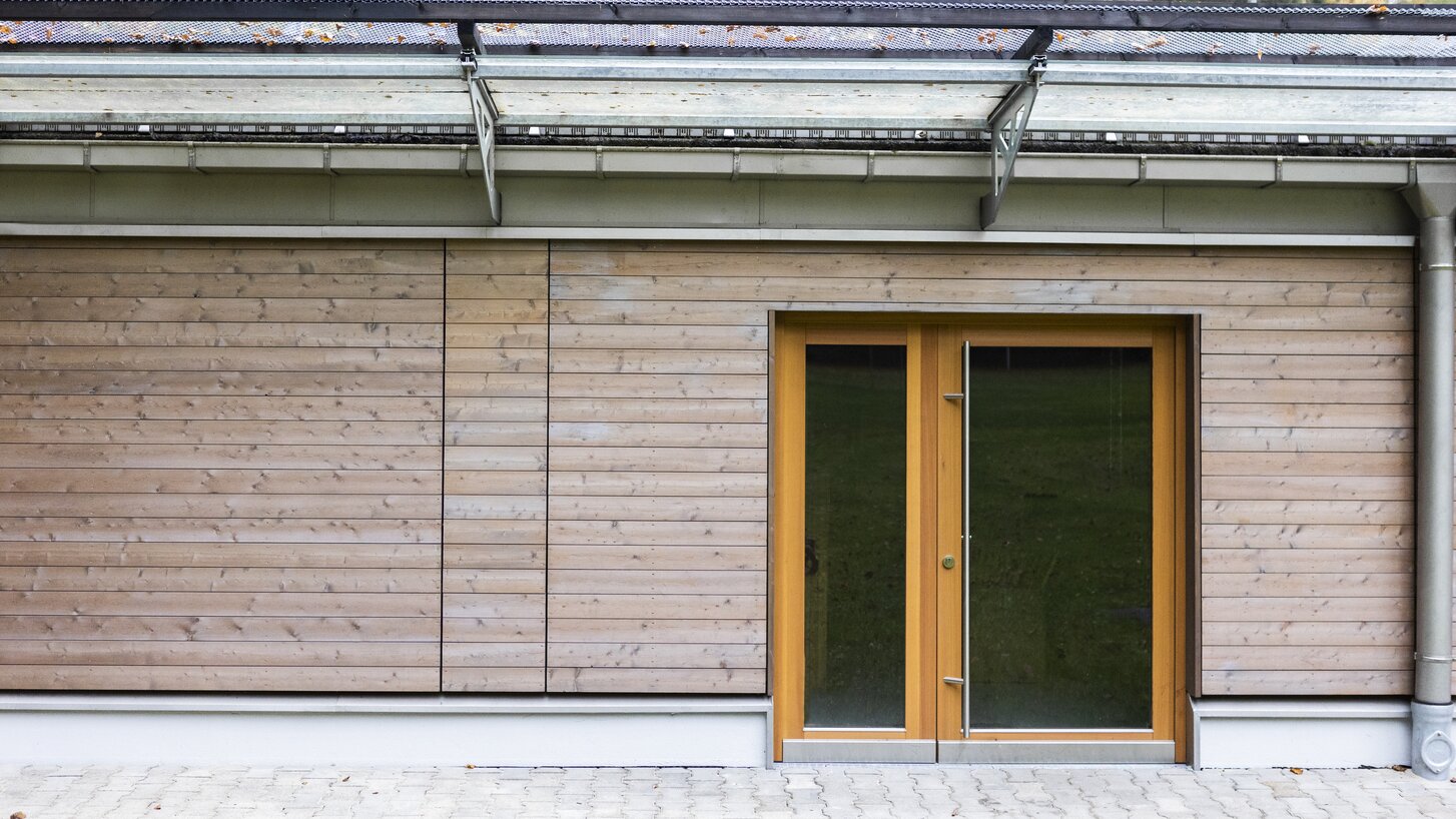 Außenwand eines Gebäudes mit einer horizontal verlaufenden Schalung aus Holz und einer Holztür | © Regina Sedlmayer