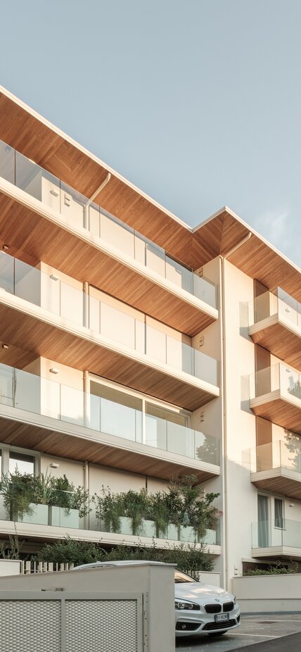 Ein mehrgeschossiges Wohngebäude mit Balkonbrüstungen aus Glas und Balkon- und Dachuntersichten aus Holz | © Davide Perbellini