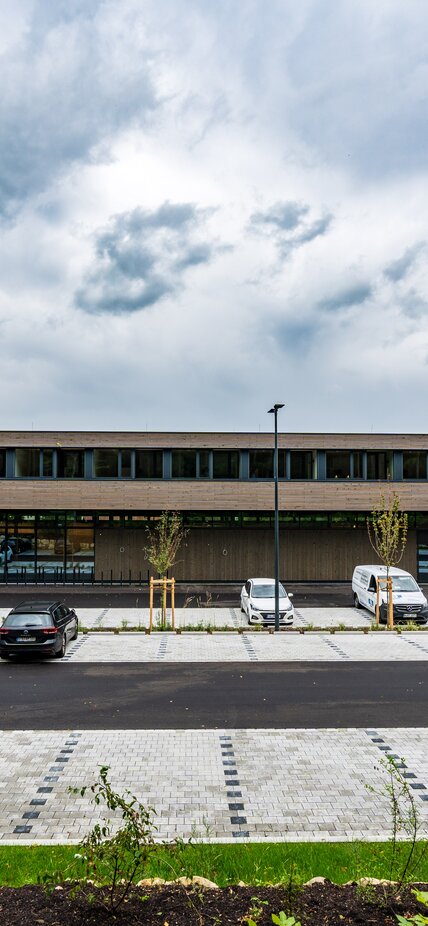 Un ampio edificio a due piani con un rivestimento di facciata in doghe di legno e un parcheggio antistante | © Roland Wehinger